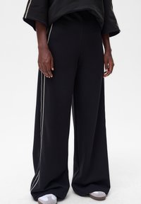 Zwarte wijde broek met een zachte textuur, voorzien van zijaccenten. Gecombineerd met grijs-witte sneakers voor een complete look.