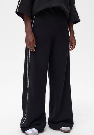 Pantalon large noir avec une texture douce, doté d'accents de passepoil sur les côtés. Assorti à des baskets grises et blanches pour un look complet.