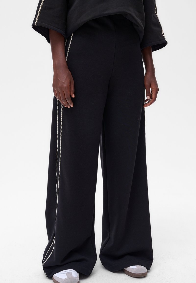 Zwarte wijde broek met een zachte textuur, voorzien van zijaccenten. Gecombineerd met grijs-witte sneakers voor een complete look.