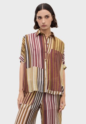 Vrouw die een loszittend shirt en broek draagt met verticale meerkleurige strepen in bruine, gele, witte en bordeauxrode tinten.