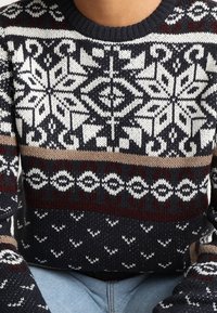 Pull-over en maille aux motifs géométriques bleu marine, blanc et beige, avec un col rond et des détails texturés sur les manches.