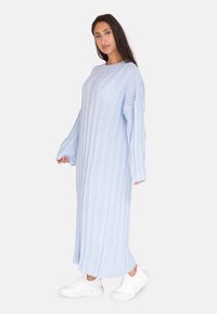 Hellblaues gestricktes Maxikleid mit vertikaler Rippstruktur, langen Ärmeln und lockerer Passform, kombiniert mit weißen Sportschuhen.