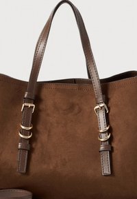 Brun ruskind shoppingtaske med to flade håndtag med guldfarvet hardware, accentstropper og en glat indvendig foring. Enkel og funktionel design.