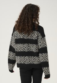 Ženska pletena majica z narebrom v črno-kremnih črtah, ki vključuje geometrijske vzorce. Ohlapen kroj, teksturiran material in rebrasti manšeti.