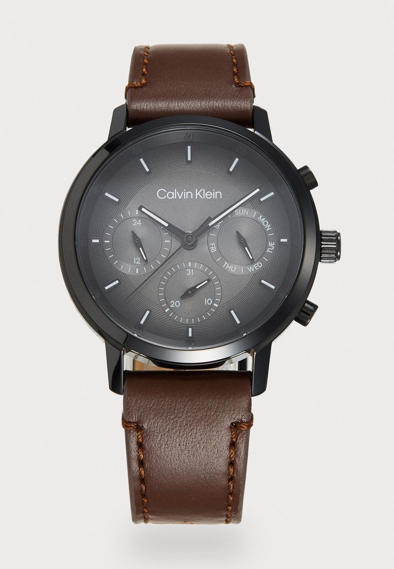 Montre Calvin Klein avec un boîtier en métal noir, un cadran gris avec deux sous-cadrans, et un bracelet en cuir marron avec des détails de couture.