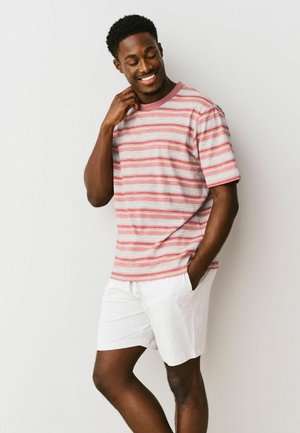 Mężczyzna uśmiechający się z zamkniętymi oczami, ubrany w różowo-białą pasiastą koszulkę i białe szorty, stojący na tle jednolitego jasnego tła.