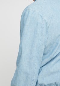 Ljusblå chambrayskjorta med knäppningkrage, långa ärmar och texturerad tyg. Synlig söm längs sömmarna.