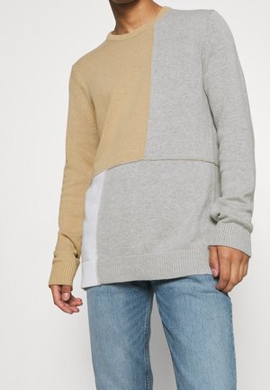 Mand iført beige, lysegrå og hvid farveblok-strikket sweater med lyseblå jeans, stående mod en ensfarvet baggrund.