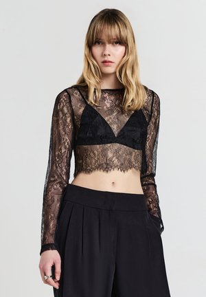 Schwarzes Spitzen-Crop-Top mit langen Ärmeln und gewelltem Saum, darüber getragen ein schwarzer Bralette. Kombiniert mit hochtaillierten schwarzen Hosen.