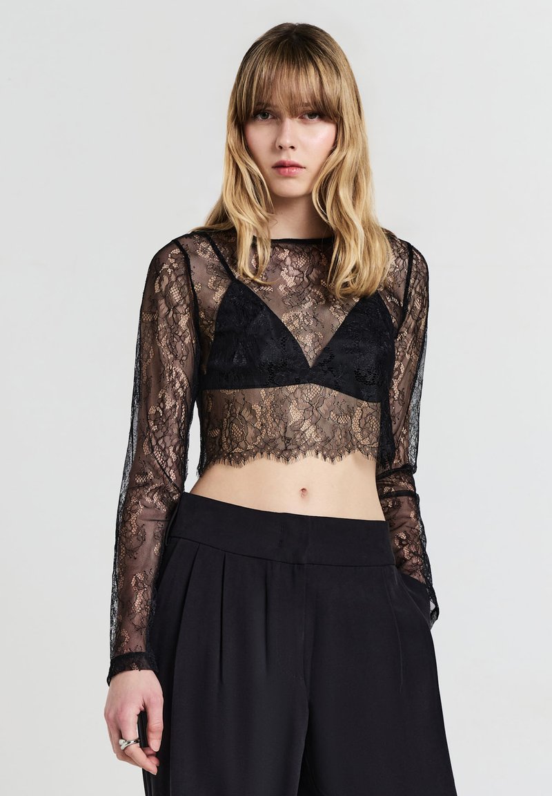 Schwarzes Spitzen-Crop-Top mit langen Ärmeln und gewelltem Saum, darüber getragen ein schwarzer Bralette. Kombiniert mit hochtaillierten schwarzen Hosen.