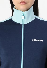 Mornarska zip-up jakna z svetlo modrimi poudarki in visokim ovratnikom. Vključuje izvezen bel logo na levi prsni strani in teksturirano tkanino.