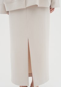 Beige midi pencilskørt med slids bagpå, båret med en matchende beige jakke, der viser dele af bærerens ben og hænder.