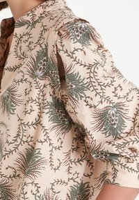 Květinová blůza s jemně růžovým pozadím, obsahující složité zelené a burgundské vzory, dlouhé rukávy a volný střih.