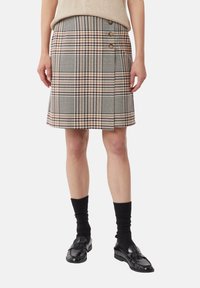 Geruite rok in beige, bruin en zwart, met een zijsplit en drie knopen in gouden tint als detail. Draagt zwart sokken en schoenen.