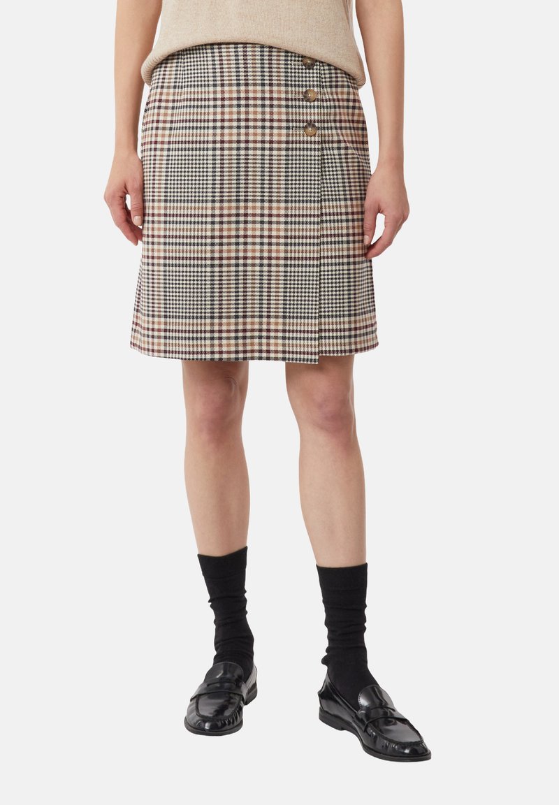 Geruite rok in beige, bruin en zwart, met een zijsplit en drie knopen in gouden tint als detail. Draagt zwart sokken en schoenen.