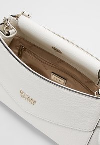 Borsa a spalla Guess in pelle bianca texturizzata con pattina aperta, interno beige, tasca con zip e dettagli in logo e hardware dorati.