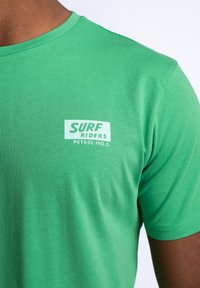 Zelené bavlněné tričko s kulatým výstřihem, s bílým logem "SURF RIDERS PETROL IND." vytištěným na levé straně hrudi. Měkká struktura.