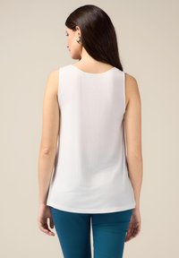 Top bianco senza maniche con scollatura rotonda, texture liscia e orlo leggermente curvo; indossato con pantaloni aderenti color teal; design minimalista.