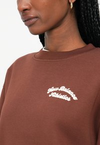 Sweatshirt castanho com decote redondo e colarinho canelado. Apresenta o logotipo branco "New Balance Athletics". Tecido de algodão com textura suave.