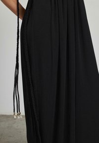 Vestido maxi negro de tejido ligero, con un panel lateral trenzado y largos flecos con acentos metálicos en el dobladillo.