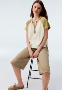 Short-sleeve, cream and olive green top with a buttoned neckline, paired with beige knee-length shorts and light brown slip-on shoes.