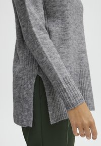 Pull en tricot gris avec un ourlet et des poignets côtelés, des fentes latérales et une texture douce, associé à un pantalon vert.