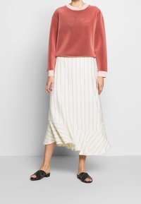 Sudadera de velour rosa con mangas largas, combinada con una falda blanca asimétrica con rayas verticales. Sandalias negras tipo slide completan el conjunto.