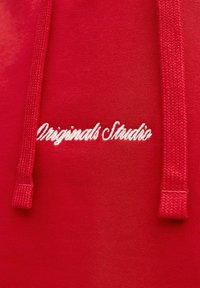 Sweatshirt rouge au toucher lisse, avec des cordons de serrage et un texte brodé blanc affichant "Originals Studio."