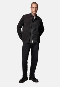 Zwarte jack met cargopockets over een donkerdenim-shirt, gecombineerd met donkere jeans en zwarte schoenen. Gladde textuur en een hedendaags ontwerp.