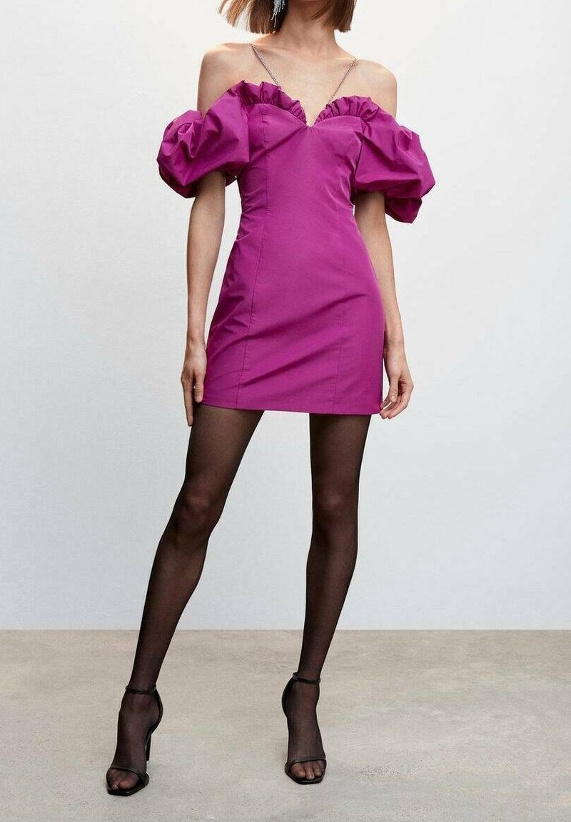 Femme portant une robe courte violette vive à épaules dénudées avec des manches bouffantes, des collants noirs transparents et des talons ouverts noirs, debout sur un sol uni.