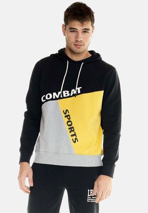 Giovane uomo che indossa una felpa con cappuccio color block nero, grigio e giallo con scritta "COMBAT SPORTS" e pantaloni della tuta neri con stampa bianca.