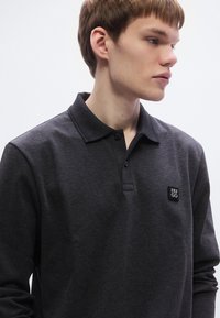 Σκούρο γκρι μακρυμάνικο πόλο πουκάμισο κατασκευασμένο από υφασμάτινη ύφανση, με γιακά με κουμπιά και μικρό επώνυμο ετικέτα στο αριστερό στήθος.