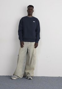 Felpa blu marina con un piccolo logo bianco, abbinata a pantaloni cargo chiari con tasche laterali e un taglio ampio. Le sneakers completano il look.