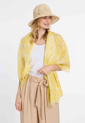 Geel bloemenpatroon sjaal gemaakt van lichtgewicht stof, draped over een witte top, gecombineerd met beige high-waisted broeken en een strohoed.