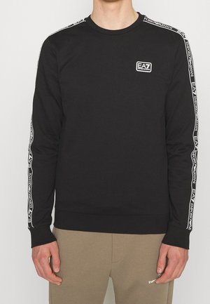 Sweat-shirt noir à manches longues avec logos blancs EA7 Emporio Armani sur la poitrine et bandes le long des manches, porté avec un pantalon beige.