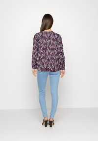 Atasan lengan panjang bermotif dalam nuansa ungu dan biru, dipadukan dengan jeans skinny biru muda dan sepatu hak tinggi hitam.