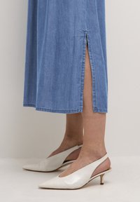 Džínové maxi šaty v světle modré barvě s bočním rozparkem. Kombinované s bílými lodičkami na podpatku se zavazováním vzadu, které mají štíhlý podpatek a texturovaný materiál.