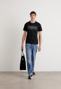 Tricou negru din bumbac cu logo-ul "BARBOUR INTERNATIONAL", asortat cu blugi albaștri deschiși și adidași negri, purtând o geantă tote neagră.