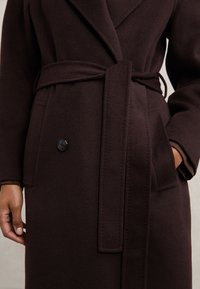 Manteau en laine brun foncé avec une large ceinture en tissu nouée à la taille, un bouton visible, des poches latérales et des manches longues.