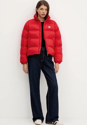 Mujer con chaqueta acolchada roja de Adidas, camiseta negra, vaqueros anchos oscuros y zapatillas blancas con rayas negras, de pie contra un fondo liso.