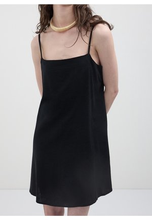 Femme portant une robe nuisette noire sans manches à fines bretelles et un collier ras du cou doré, debout devant un arrière-plan gris clair.