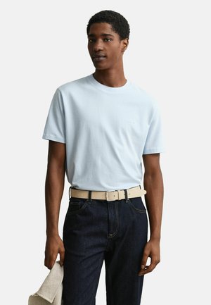 Mann trägt hellblaues kurzärmeliges T-Shirt, in dunkle Jeans mit beigem Gürtel gesteckt, hält einen beigen, strukturierten Stoff in der linken Hand.