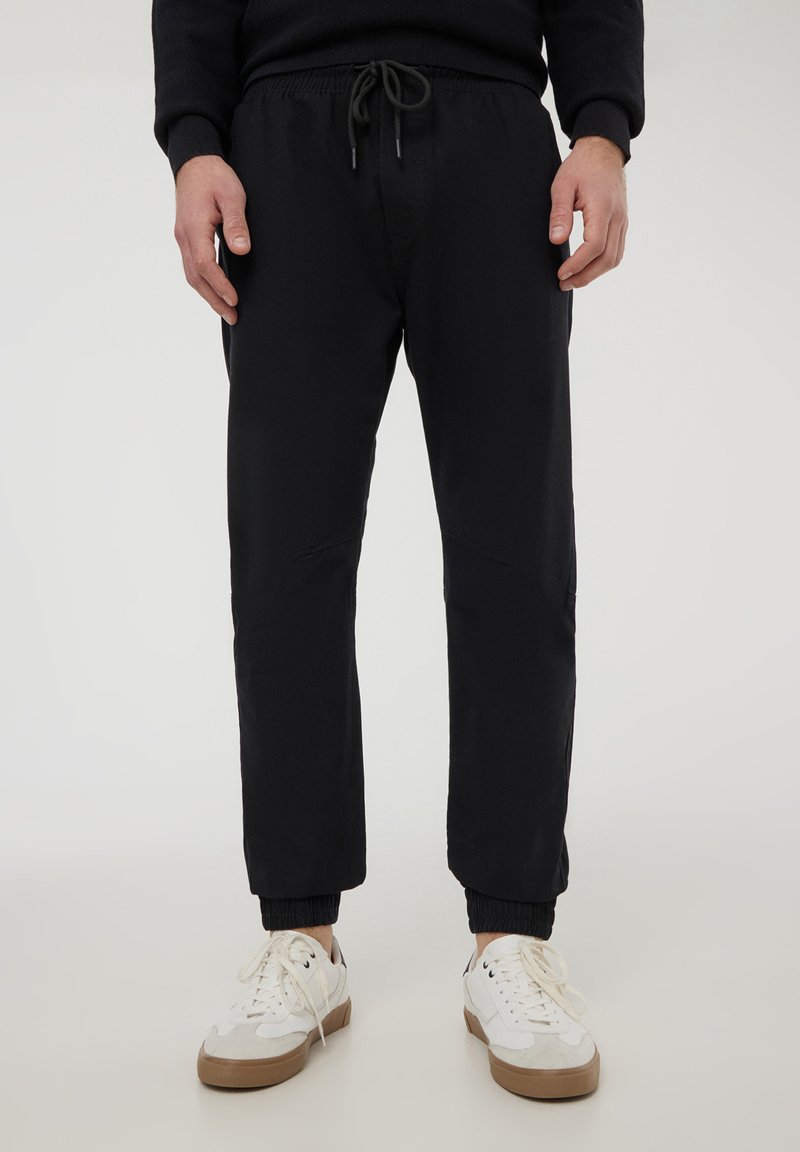Pantaloni jogger neri realizzati in tessuto morbido, dotati di vita elasticizzata con cordoncino, polsi a costine e dettagli di cucitura sulle ginocchia.