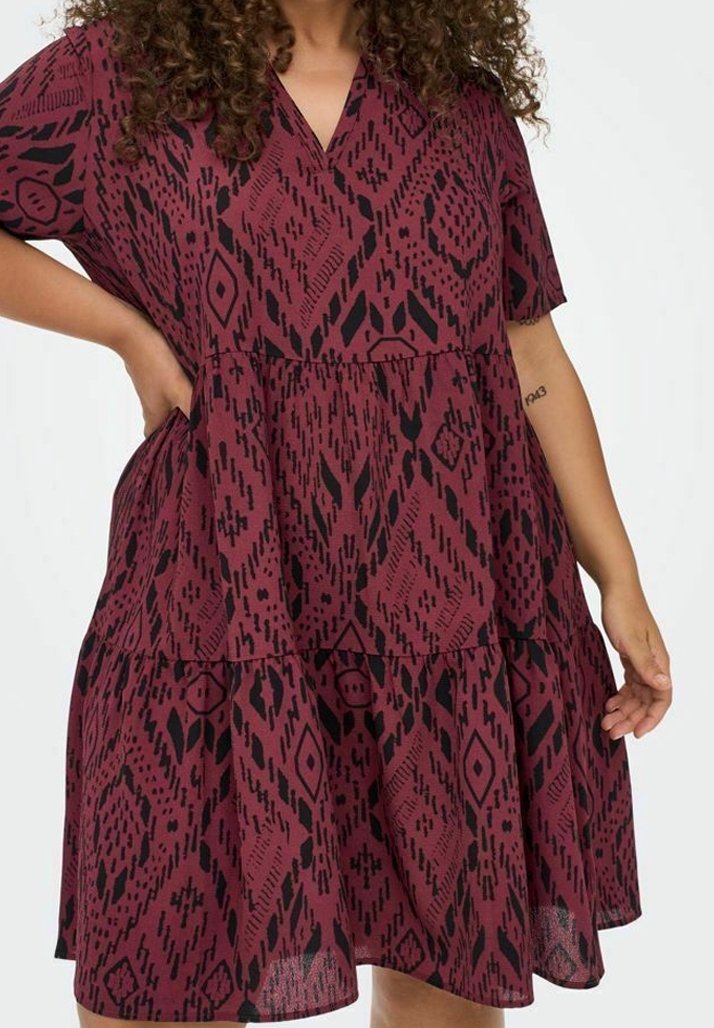 Femme portant une robe ample bordeaux à manches courtes avec des motifs géométriques noirs, un col en V et une jupe à volants.