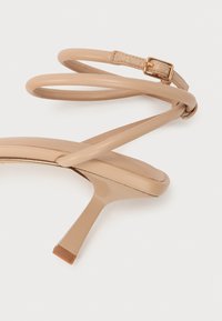 Sandale à talons hauts beige avec finition brillante, fines lanières enroulées autour de la cheville et bout carré. Détail de boucle ajustable sur la lanière.