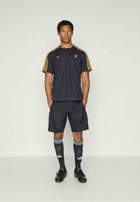 Črna atletska majica s črtami in rumenimi poudarki, kombinirana s črnimi kratkimi hlačami in nogavicami do kolen, ki imajo bele črte in logotip. Črne nogometne čevlje.