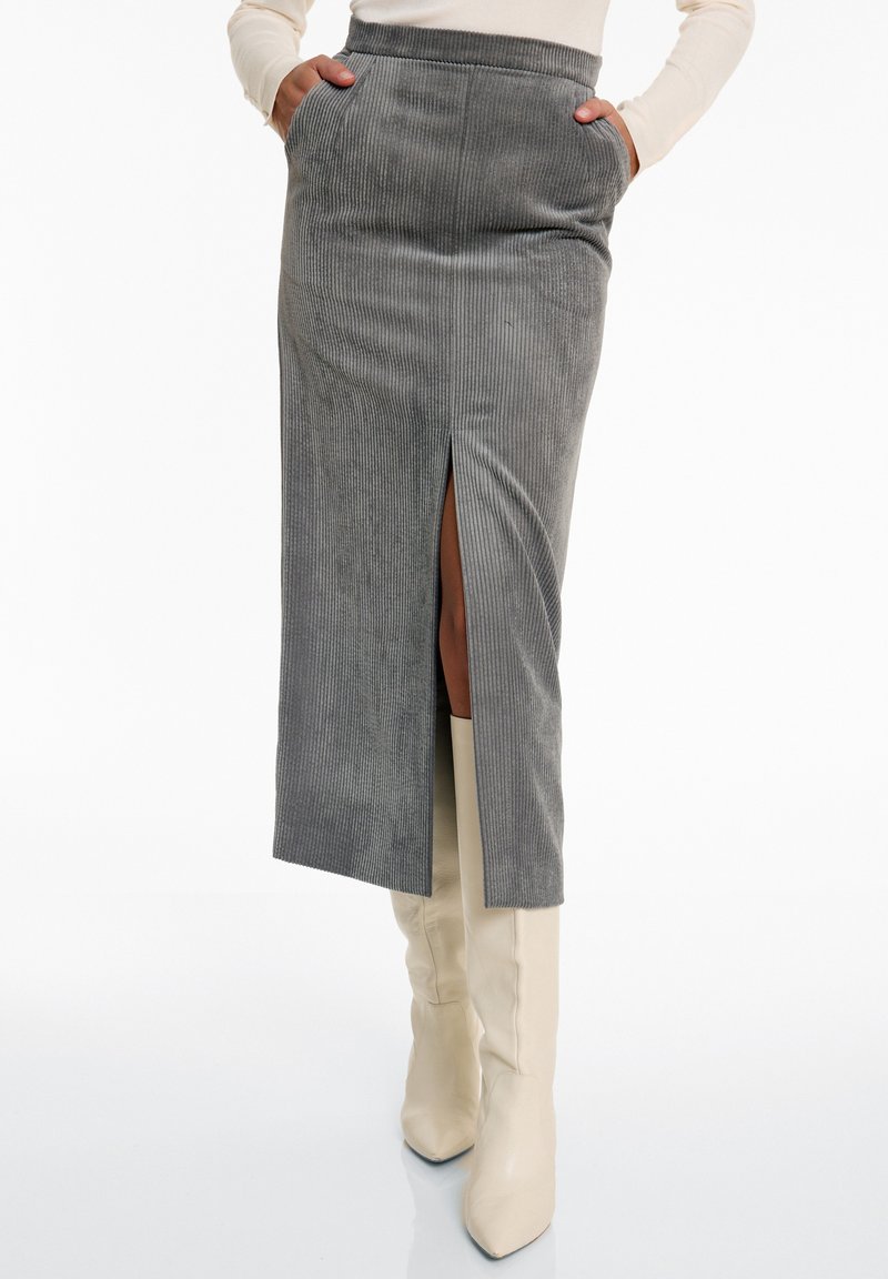 Grauer Cord-Midi-Rock mit seitlichem Schlitz, verfügt über Fronttaschen und einen taillierten Bund. Kombiniert mit knielangen cremefarbenen Stiefeln.
