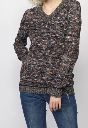 Personne portant un pull en maille multicolore à manches longues et un jean bleu, main dans la poche, sur un fond clair uni.