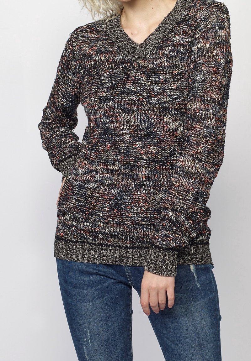 Personne portant un pull en maille multicolore à manches longues et un jean bleu, main dans la poche, sur un fond clair uni.