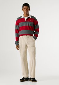 Sarkans un pelēks svītrots garās piedurknes polo krekls ar baltu apkakli, kombinēts ar krēmkrāsas kordura biksēm un melniem sporta apaviem ar baltām akcentiem.